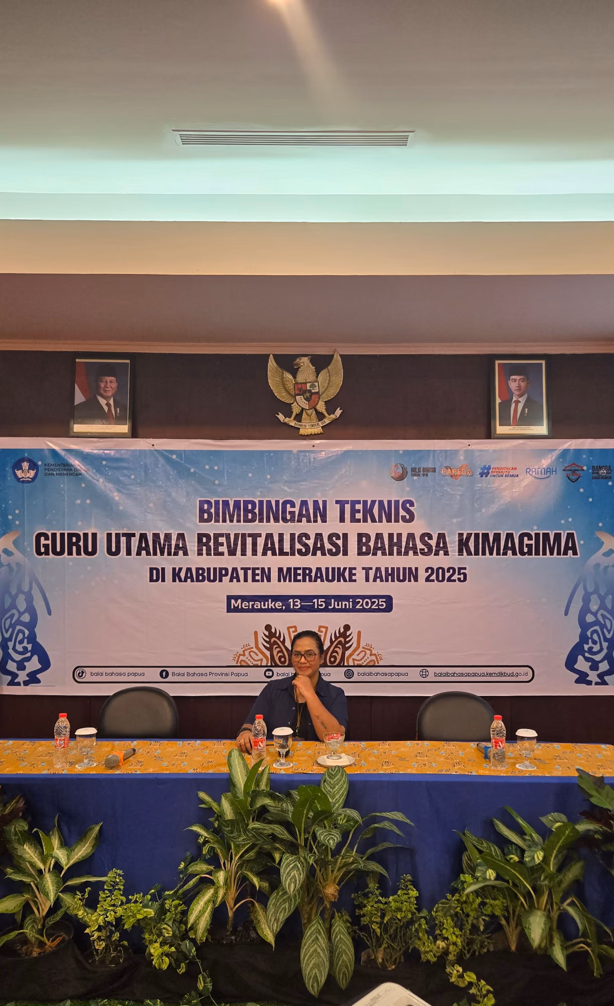 Meja panel tertata di depan backdrop kegiatan pemerintah daerah di dalam ruangan.
