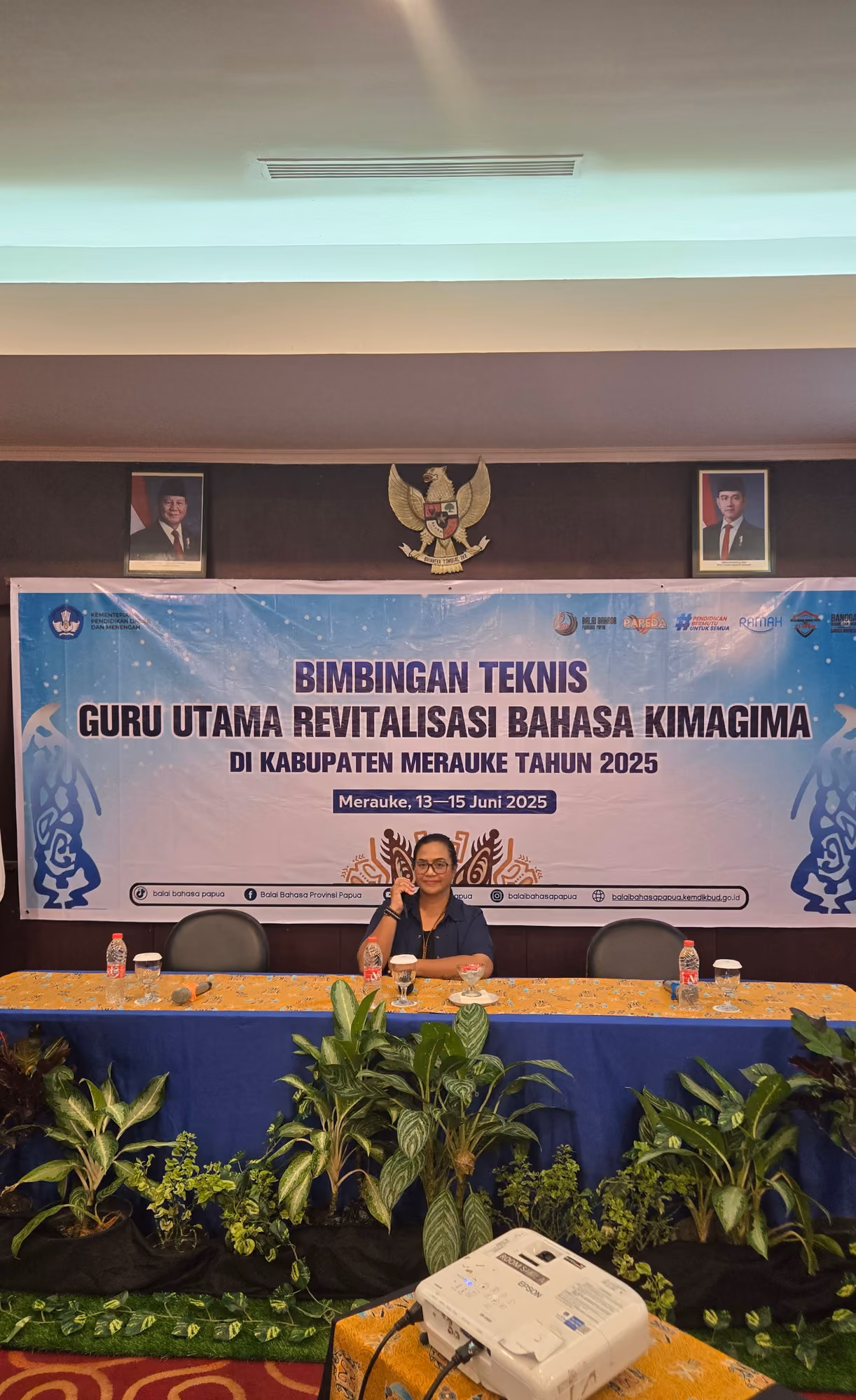 Panggung kegiatan pemerintah dengan backdrop, meja panel, dan tanaman hias.