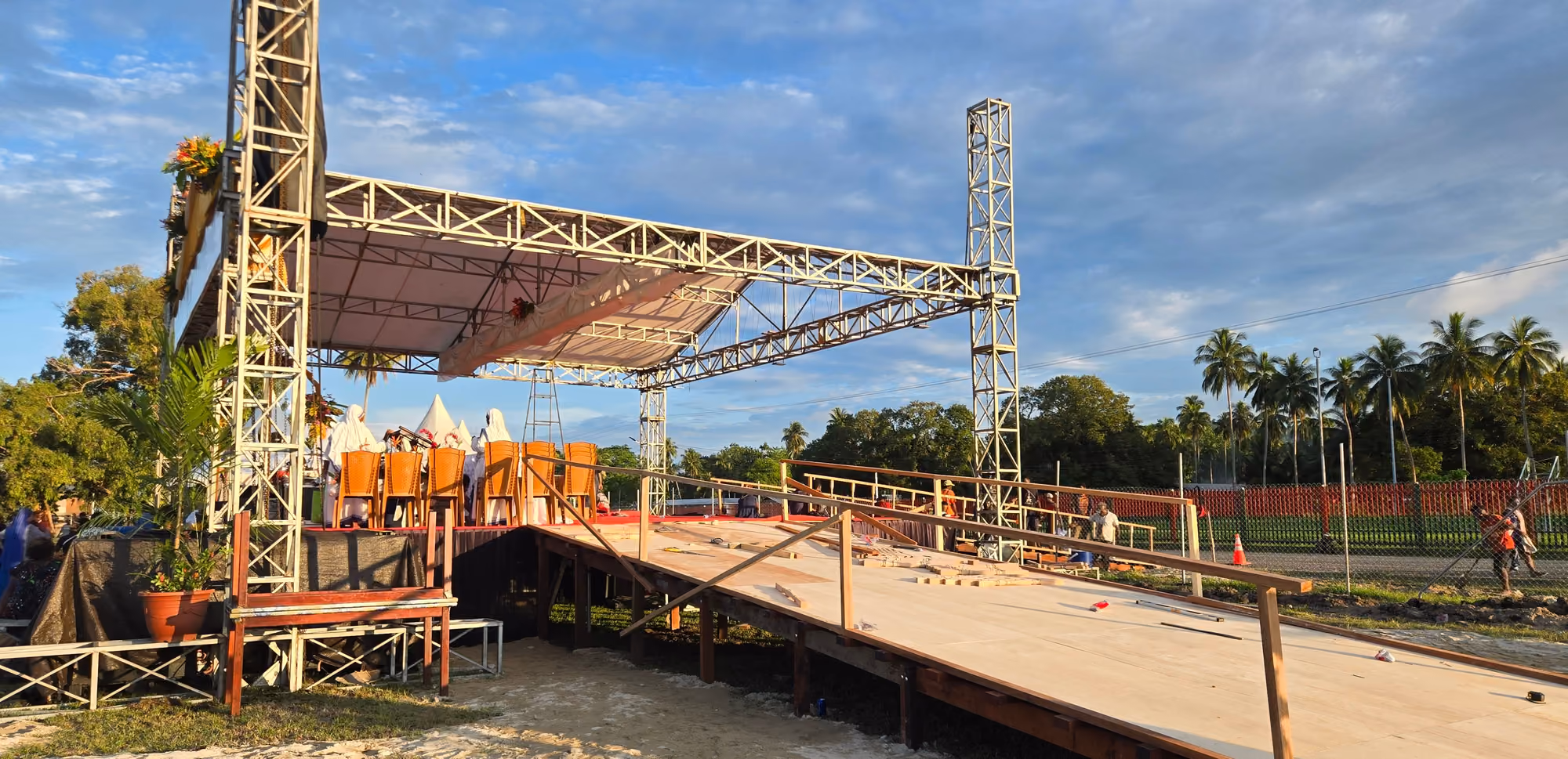Rangka baja panggung dan lantai kayu dalam proses pemasangan di lapangan terbuka pada siang hari.