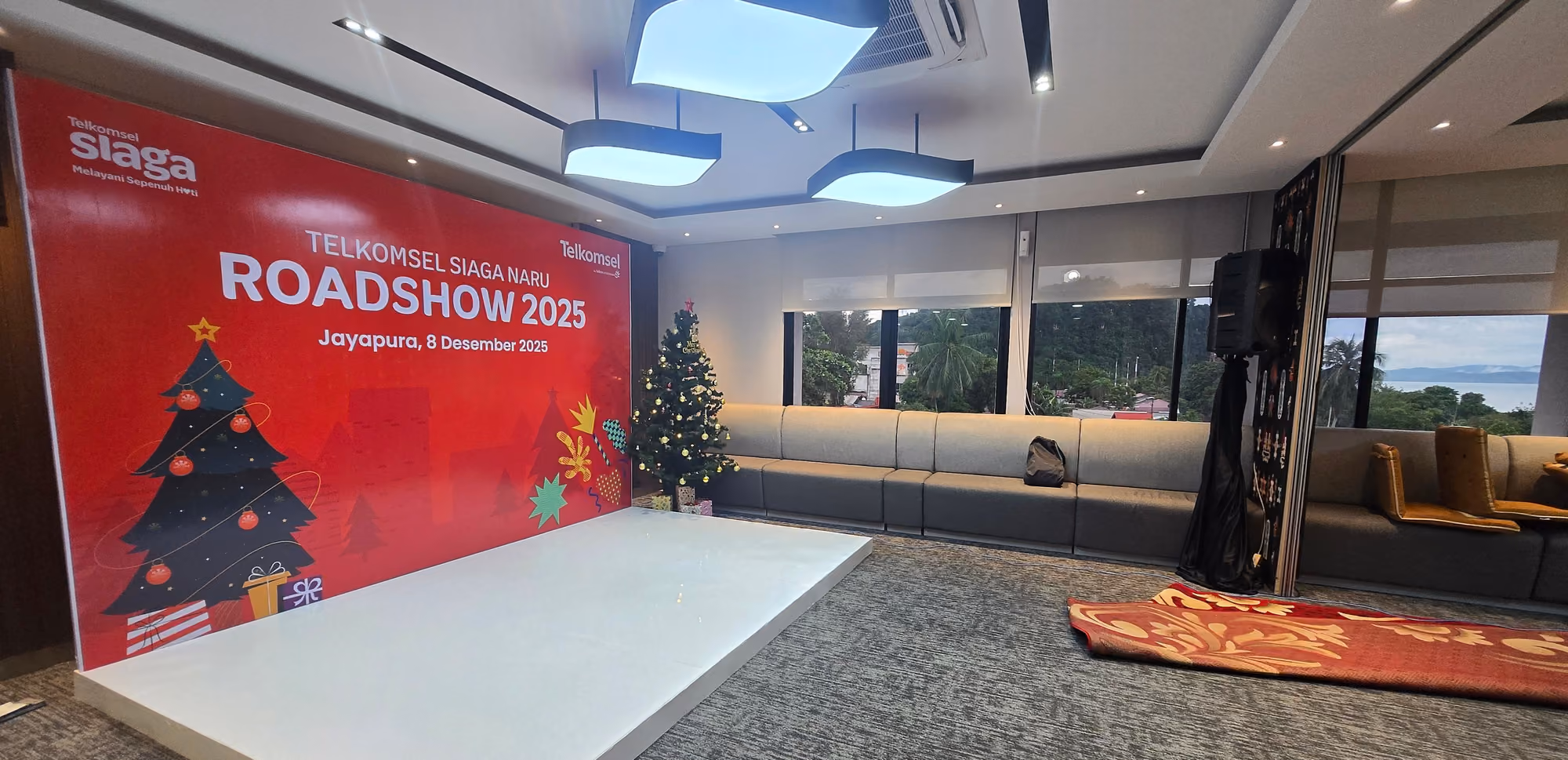 Ruang acara dalam gedung dengan backdrop merah dan panggung rendah yang telah tertata.