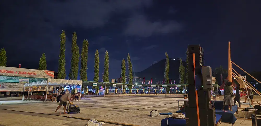 Area produksi terbuka pada malam hari dengan susunan speaker, kabel, tenda, dan kru yang menata perlengkapan acara.