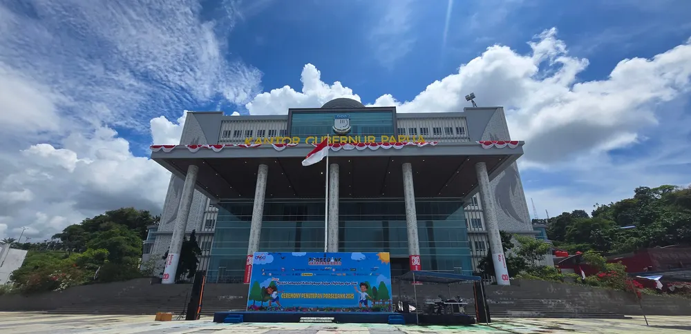 Area upacara di halaman gedung berpilar dengan panggung dan backdrop terpasang.