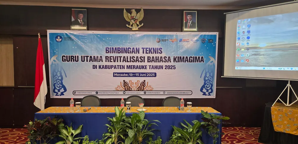 Backdrop kegiatan bimbingan teknis dengan meja panel berpelapis biru dan tanaman hias di depannya.