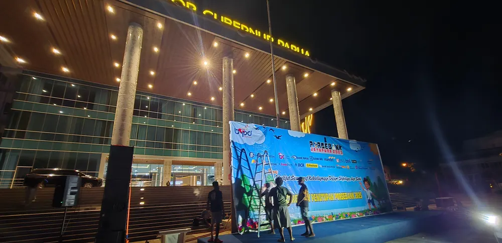 Backdrop acara terpasang di halaman gedung berpilar dengan kru menyelesaikan pemasangan.