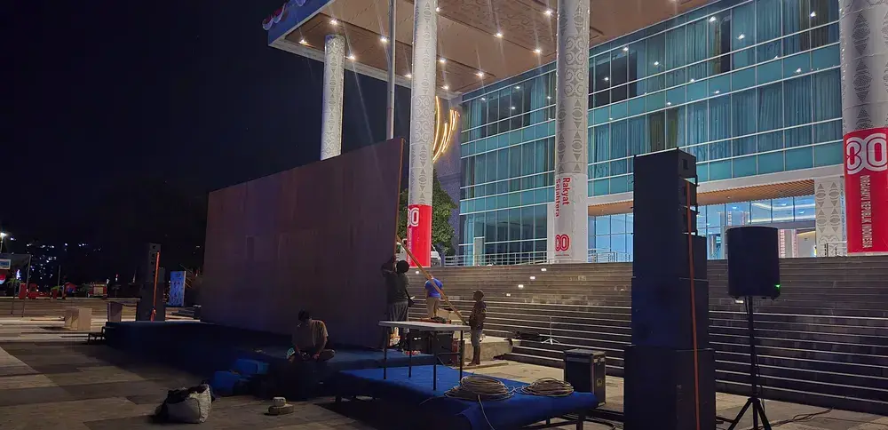 Panggung rendah, backdrop besar, dan susunan speaker sedang dipersiapkan di area plaza terbuka pada malam hari.