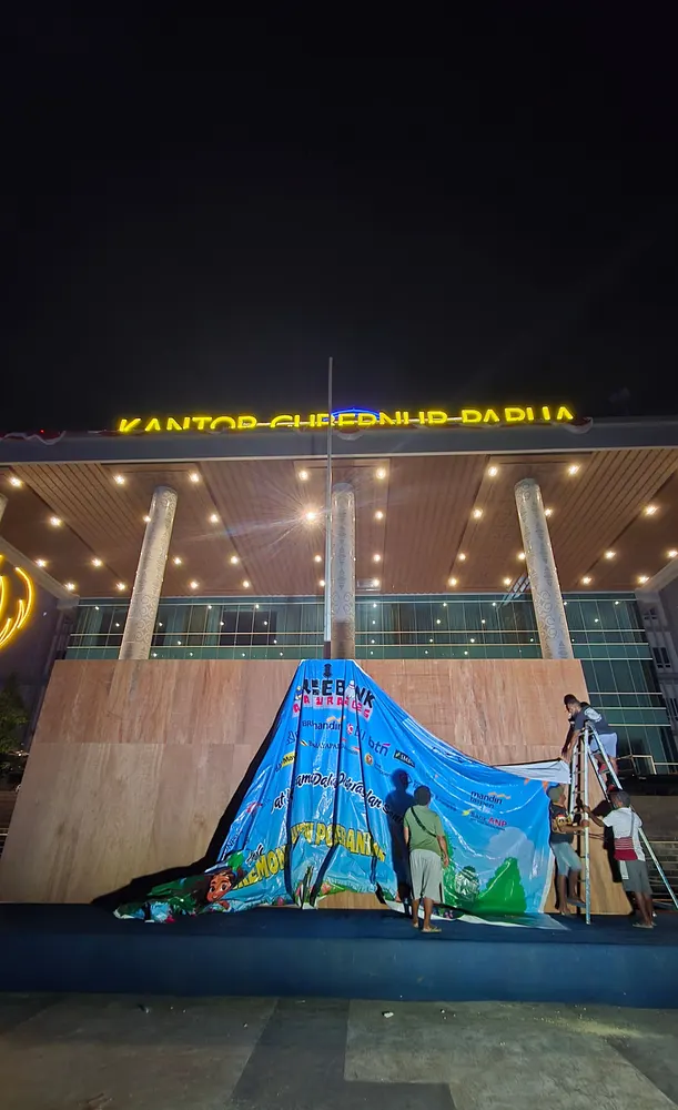 Kru memasang backdrop berukuran besar di depan gedung berpilar tinggi pada malam hari.