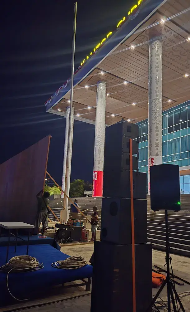 Susunan speaker produksi berdiri di samping panggung rendah dengan kabel dan kru yang sedang bekerja pada malam hari.