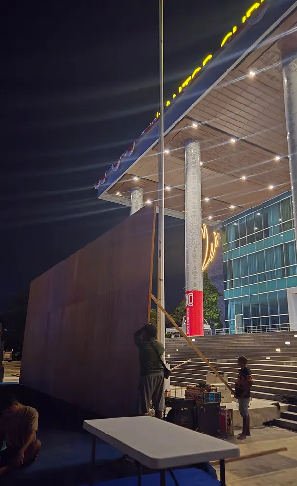Struktur dinding backdrop berdiri di area plaza terbuka menjelang malam.